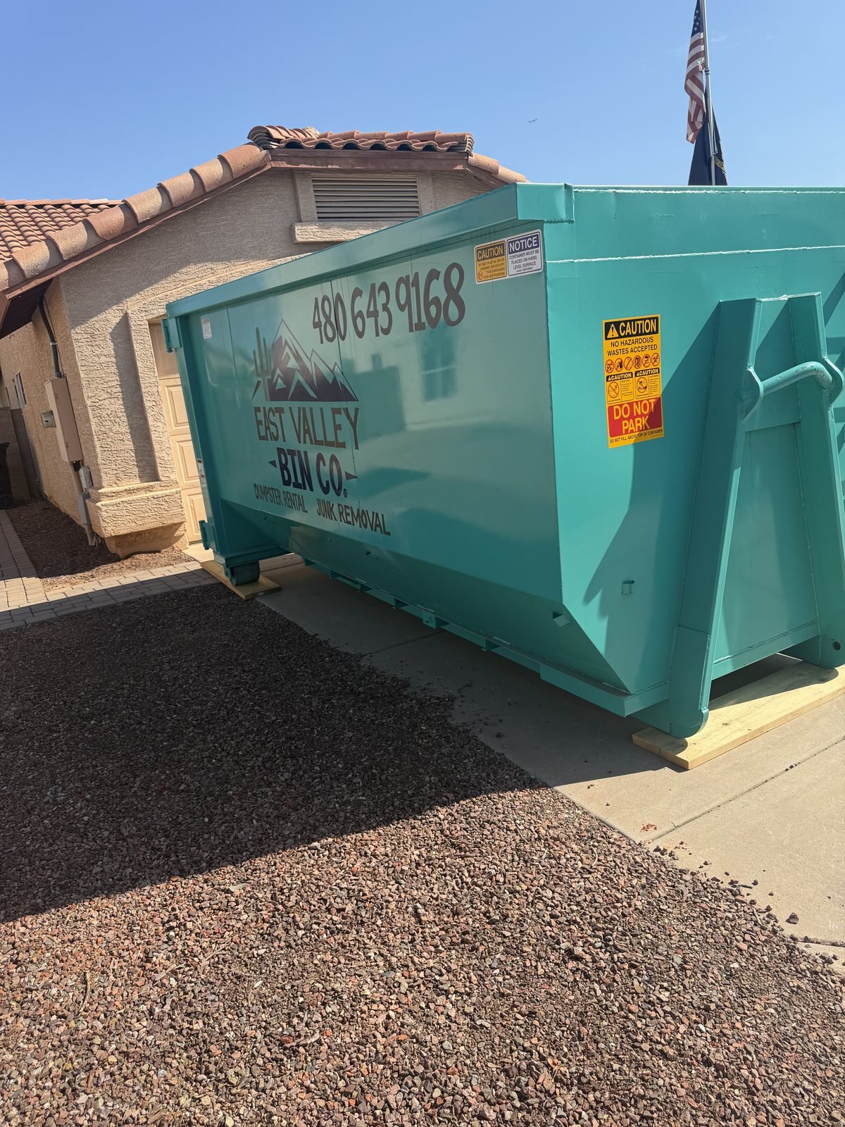 Branded teal East Valley Bin Co. dumpster set on wood protection boards on a driveway in front of a home