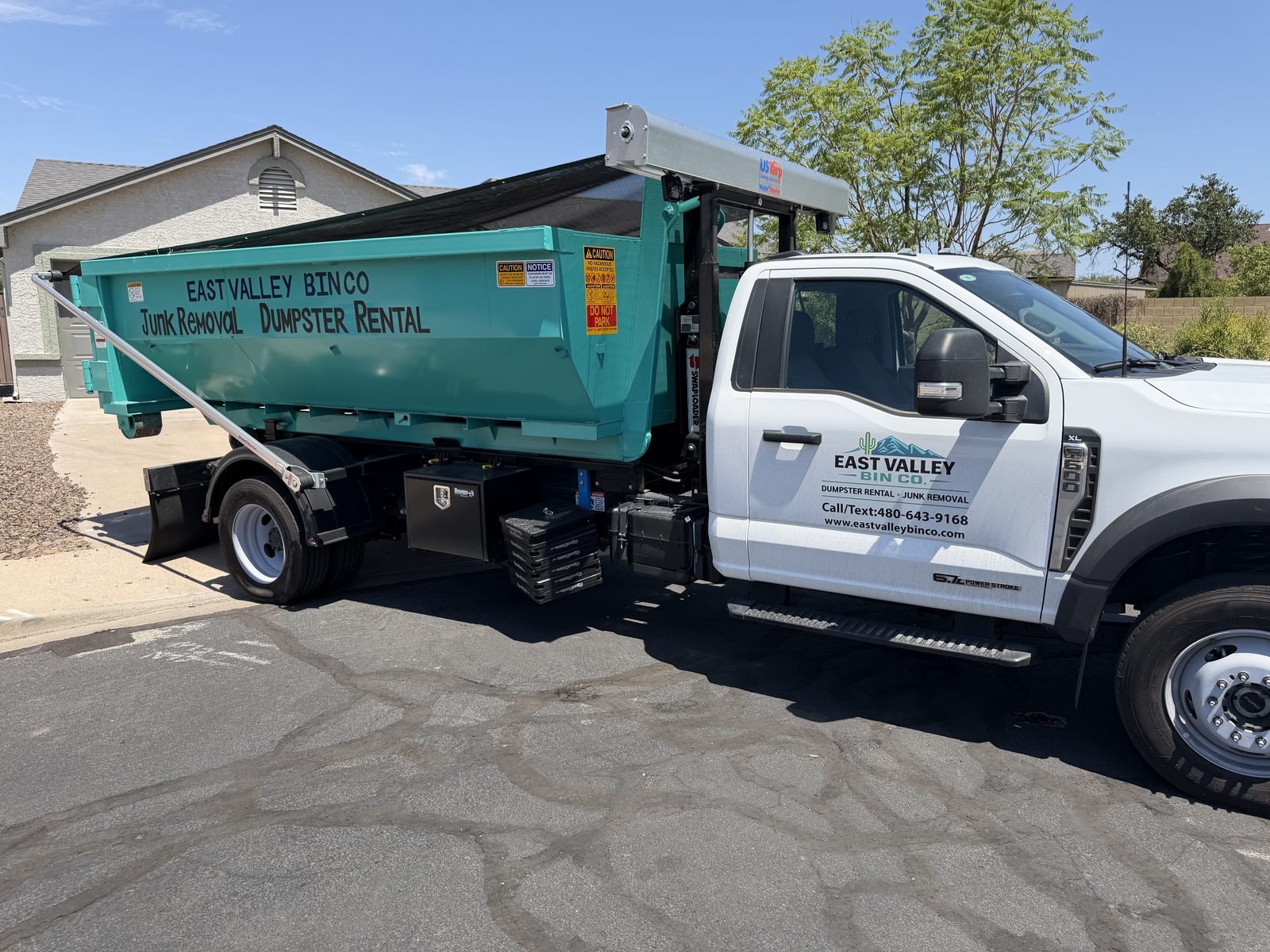East Valley Bin Co. branded roll-off truck delivering a teal East Valley Bin Co. dumpster at a home in the East Valley, Arizona